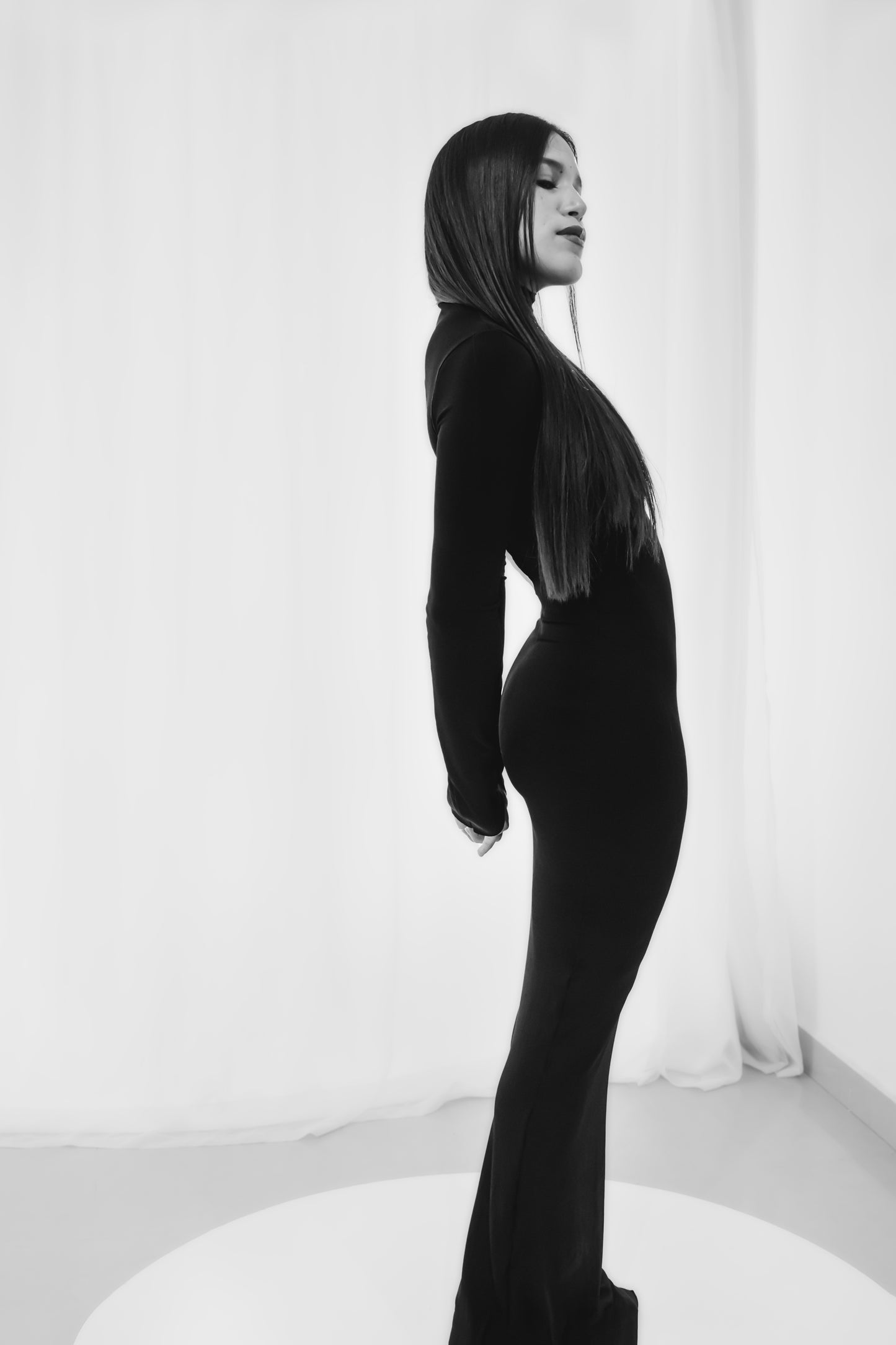Una imagen monocromática de una mujer parada de perfil usando un vestido negro ajustado con mangas largas y dobladillo hasta el suelo. La persona tiene el pelo largo y liso y posa sobre un fondo blanco.