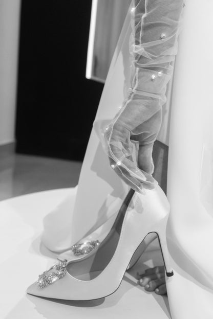 Imagen en blanco y negro que muestra el pie de una mujer calzándose un zapato blanco de tacón alto con un broche brillante, el dobladillo de su vestido y unos guantes de tul con pequeños cristales.