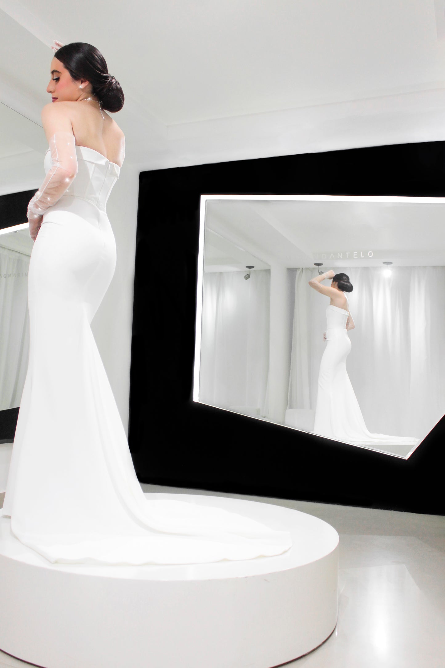 Fotografía de una mujer que lleva un vestido de fiesta blanco con una cola larga y unos guantes largos de tul con pequeños cristales, se encuentra sobre un pedestal circular blanco en una habitación bien iluminada, con su reflejo visible en un gran espejo colocado en ángulo hacia la derecha.