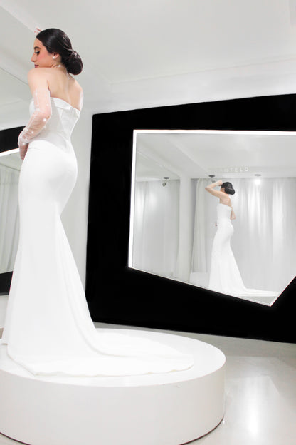Fotografía de una mujer que lleva un vestido de fiesta blanco con una cola larga y unos guantes largos de tul con pequeños cristales, se encuentra sobre un pedestal circular blanco en una habitación bien iluminada, con su reflejo visible en un gran espejo colocado en ángulo hacia la derecha.